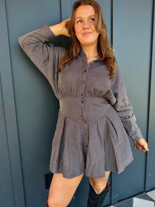 A woman wearing a gray corduroy long-sleeve mini dress with a button-down front, standing against a dark gray paneled wall.